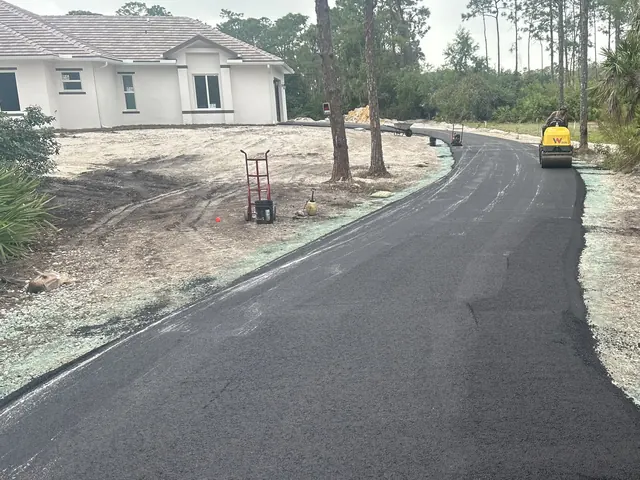 New asphalt driveway curving through sand and slash pines toward a white stucco house, with a roller at the far end