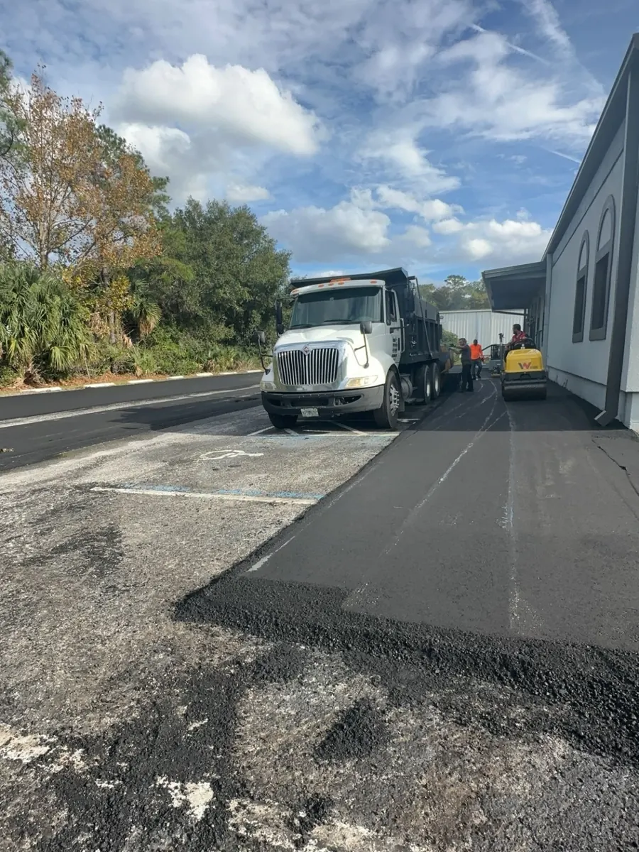 A fresh lane of asphalt laid over an old, worn parking lot, with a dump truck and roller beside a building