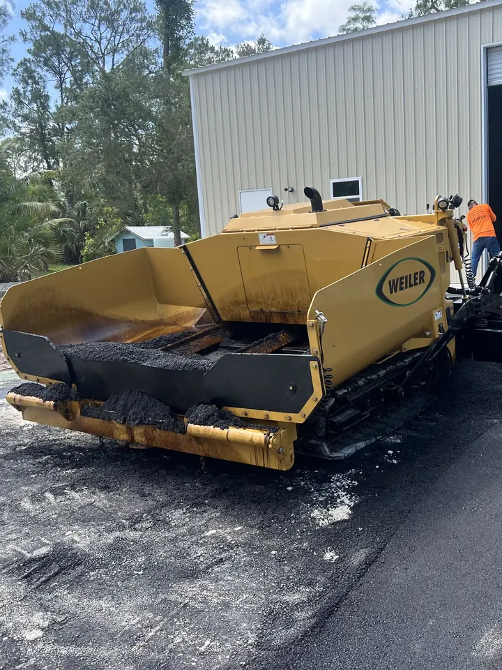Asphalt paver parked on a fresh mat in front of a large metal building with its door open