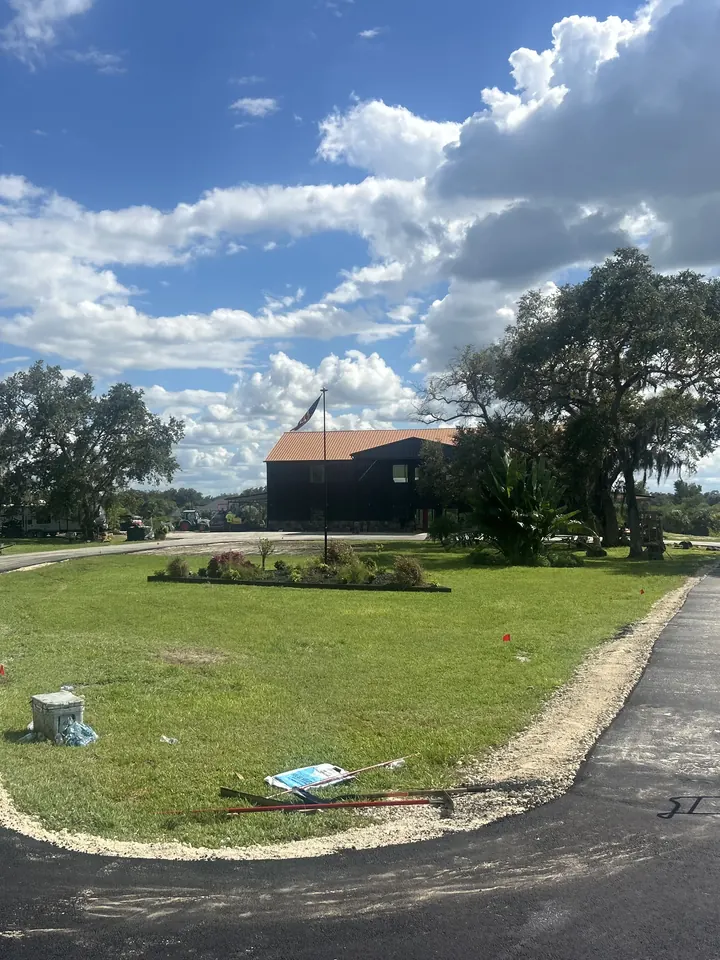 Freshly paved driveway curving around a lawn toward a dark-sided house under live oaks and a big blue sky